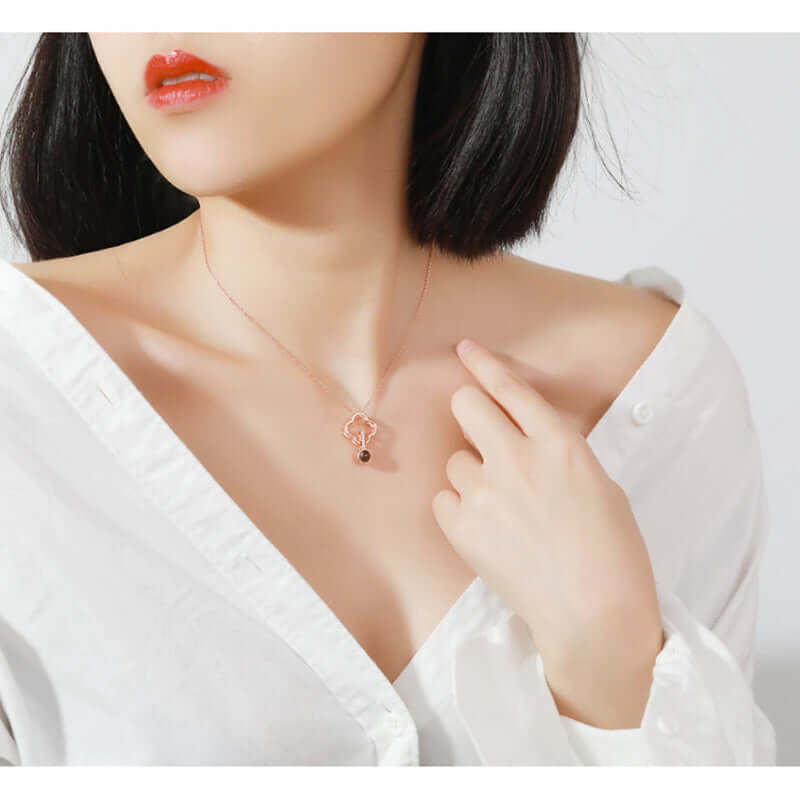 A pointing at the I love you necklace while looking right side of the picture. The women is wearing a white top and a red lipstick. 
